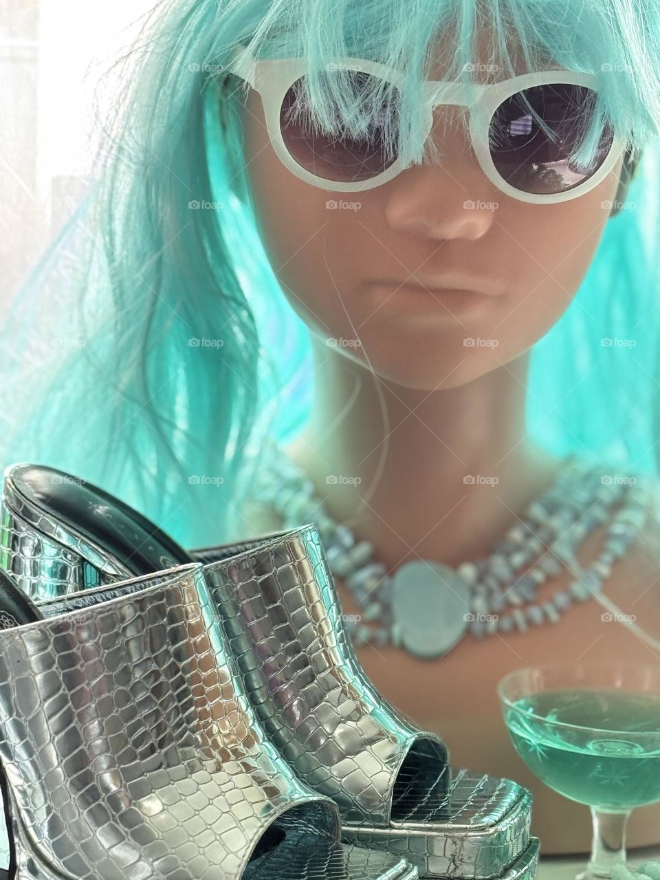 Mannequin wearing aqua wig and bead necklace with coupe glass of aqua liquid and foil alligator platform shoes 