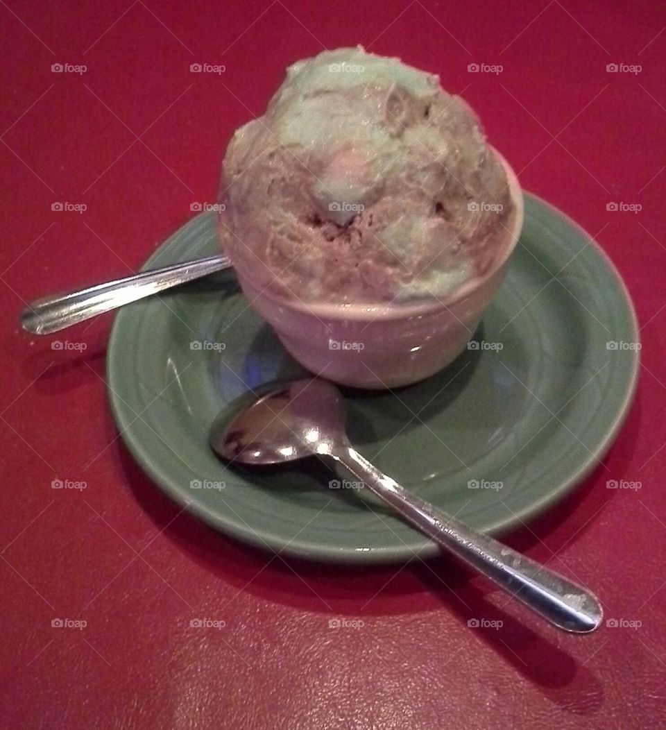 italian dessert dish, spumoni (cherry pink, green pistachio and brown chocolate) ice cream. red background and served in a clear glass dish on a green ceramic plate with 2 silver spoons for sharing.