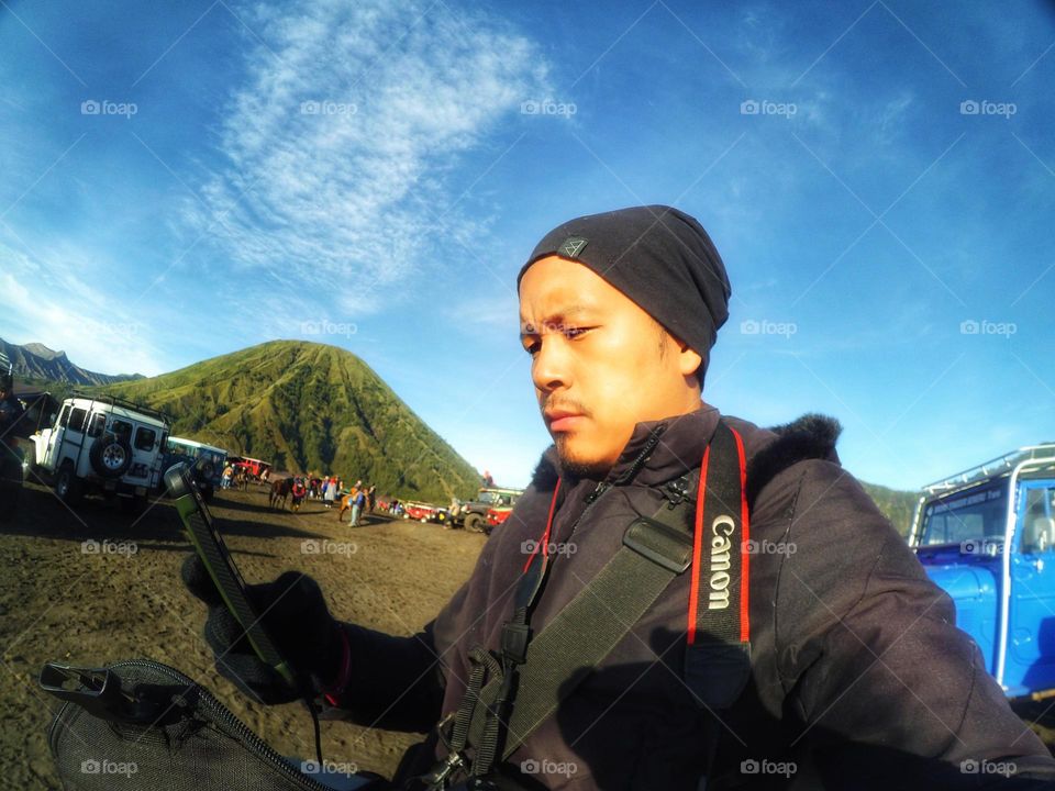 Selfie photo of a young Asian man in warm clothing against a green background in the Bromo mountains area with a cloudy sky. Vacation and adventure.