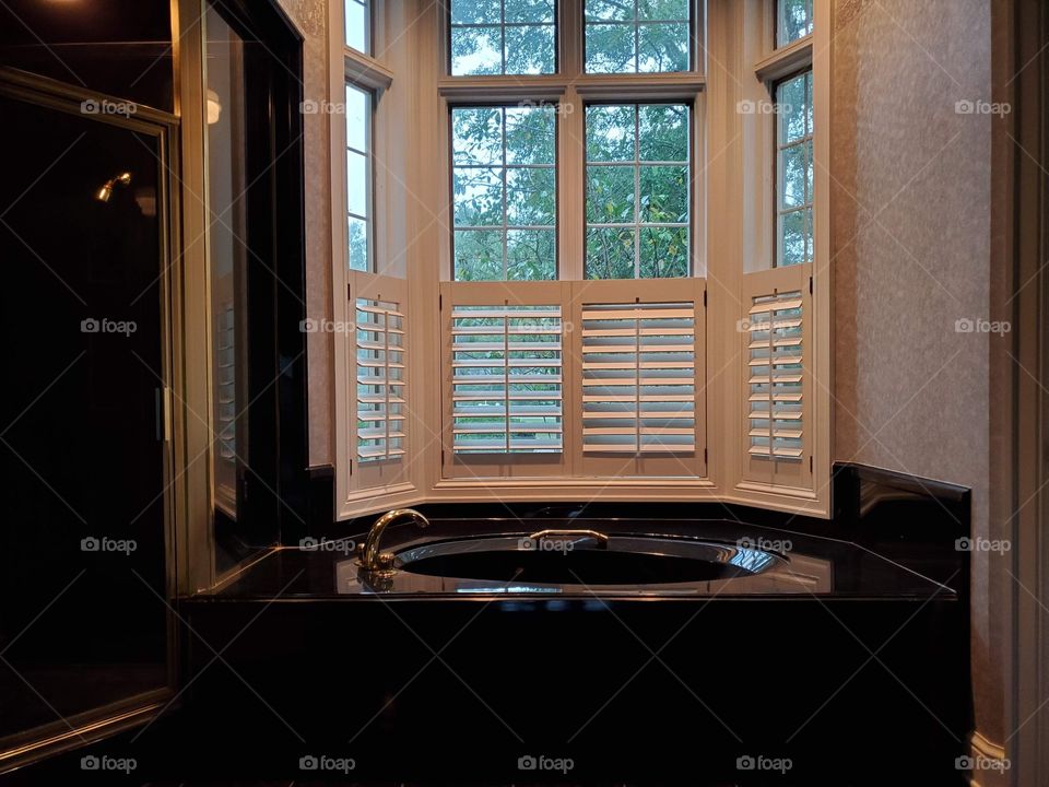 Black marble bathtub underneath a window with white slat shutters