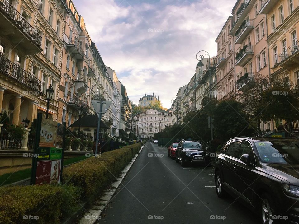 
A street with some cars parking, at the end of the street there is a splendid building, prominent between 2 blocks of street houses . cloudy sky and beautiful scenery with golden time 