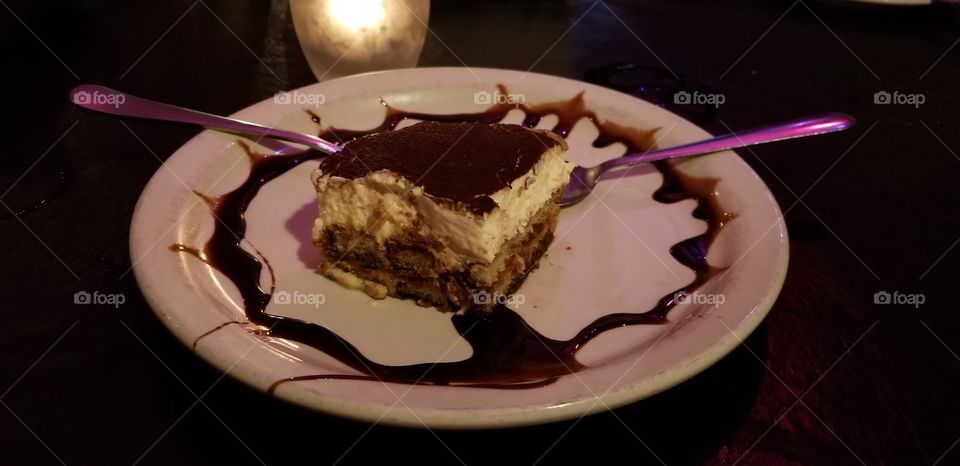A delicious of piece of tiramisu cake with chocolate sauce around it and two forks stuck inside.