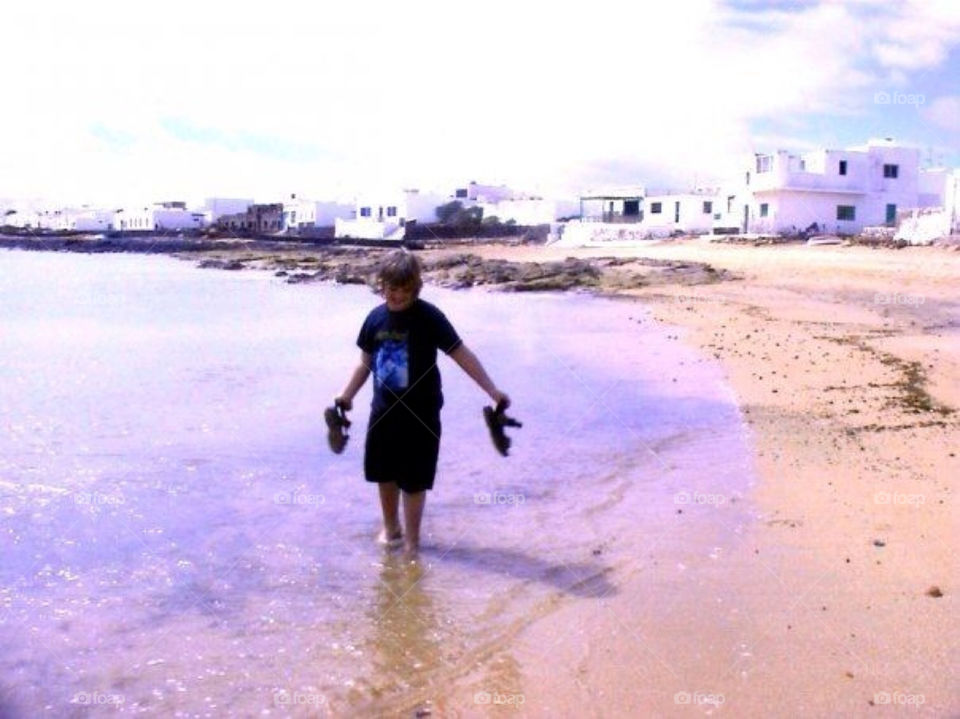 A boy walking on the beach. A boy walking in the water and holding his shoes 
