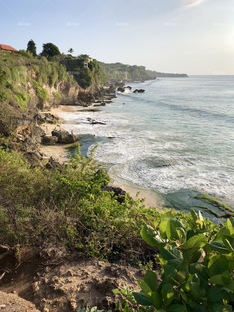 Curvy island coast. Small ocean waves. Plants in foreground. Developing country. Bali, Indonesia. 2018.