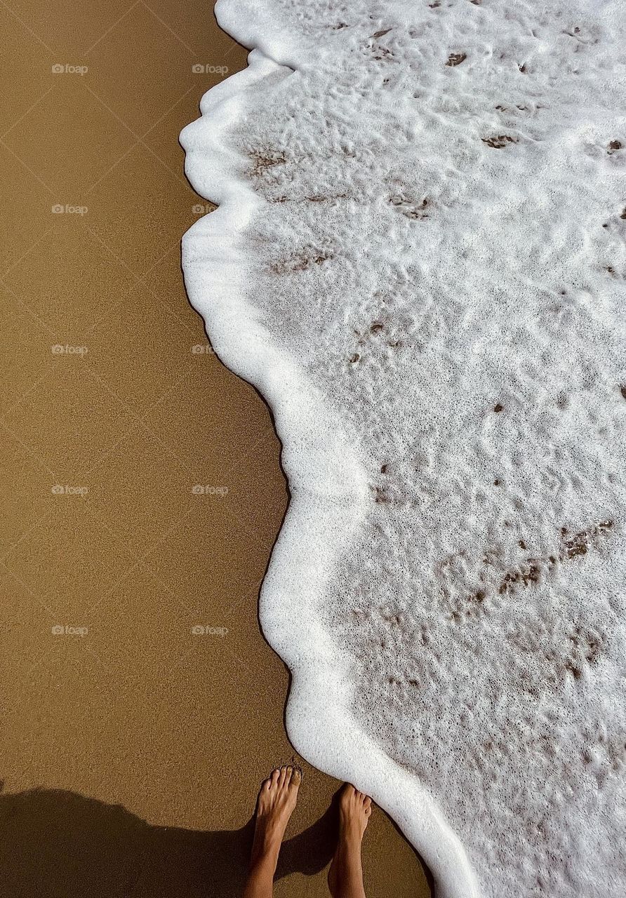 Foamy sea wave covering female feet, washing up on the Mediterranean sandy beach shore 