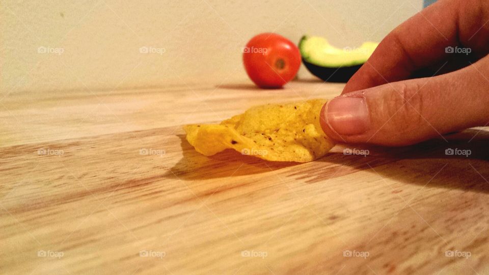 fingers holding tortilla chip with avocado and tomato in background