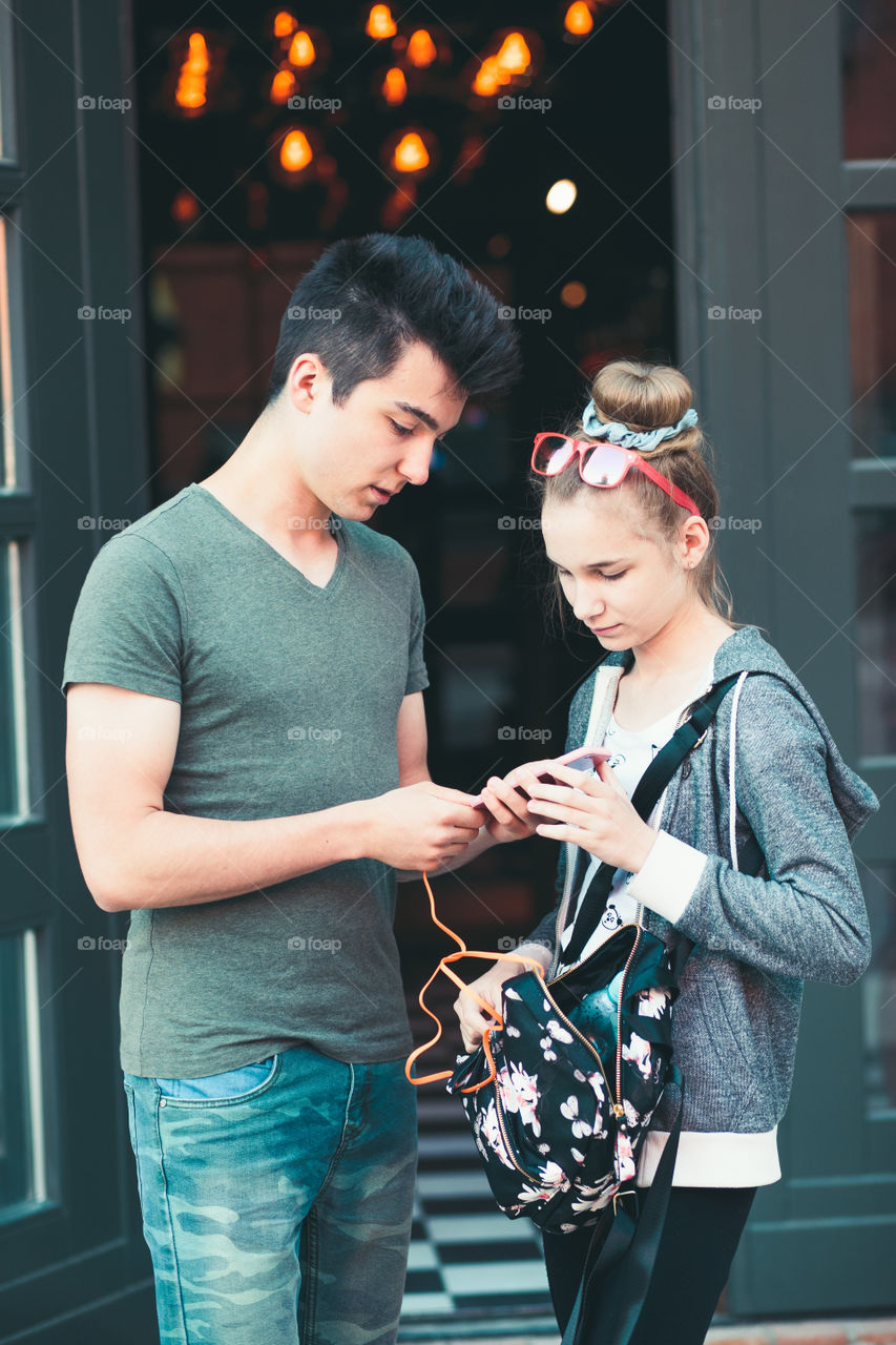 Couple of friends, teenage girl and boy, using smartphones, talking together, standing on street in center of town, spending time together