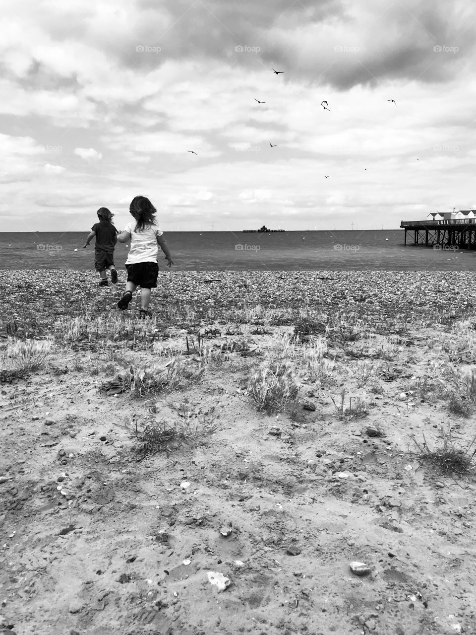 Siblings running towards the shore on pebbled beach in monochrome