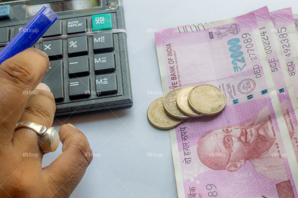 Cropped hand of a woman checking account with calculator and holding a pen. Indian currency notes and coins are on the side of table. Corporate business concept.