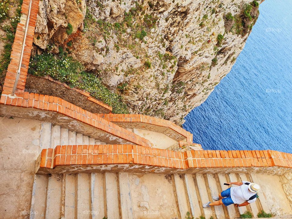 man with hat photographed from above descends the steps to the sea to reach the caves of Neptune in Sardinia