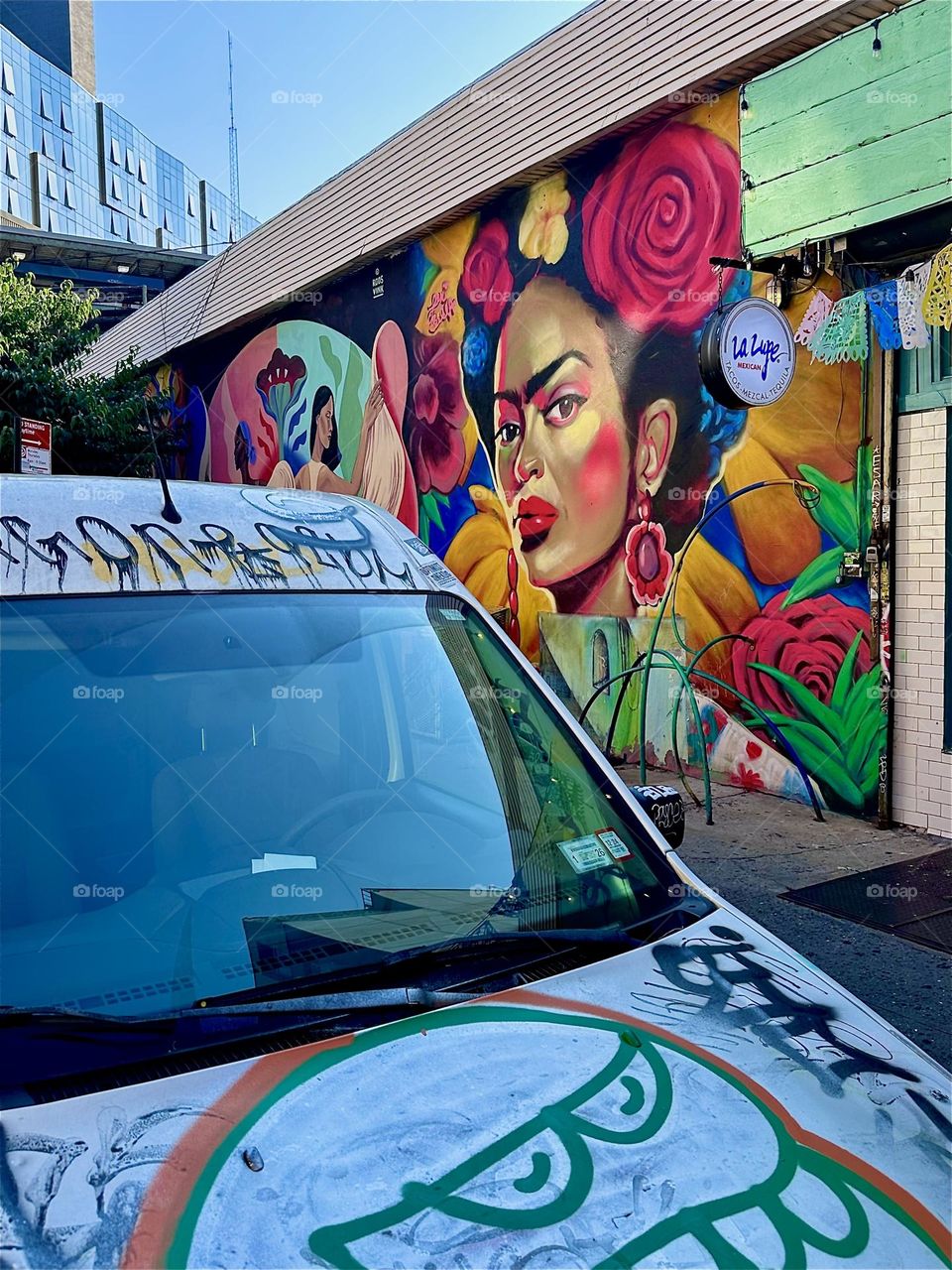 Next to a car with graffiti on “Jefferson St” off “Broadway” in “Bushwick”, Bklyn we see a large art mural celebrating the Latin culture featuring a Latina with a big red rose in her hair. The “M” train overpass is nearby. 2024. Hypnotic Productions