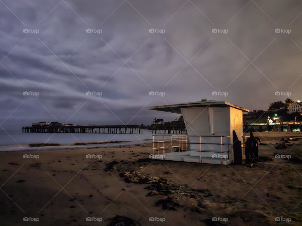 Looking down the beach at Capitola by the Sea in California at night with the lifeguard station with a soft cloudy sky, in the distance is the wharf that sustained damage in the January 2023 storms that ravaged the coastal region