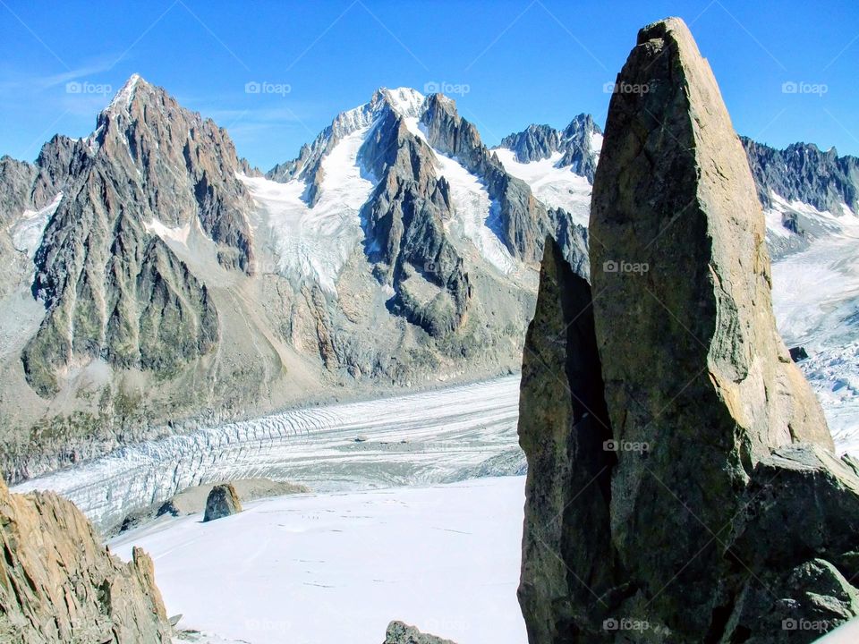 Argentiere glacier with large jagged rock formation in the foreground, mountains in the background with bright blue sky