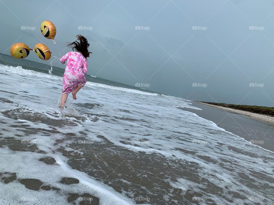 Little Girl Jumping And Running Having Fun With Helium Emoji’s Balloons On The Beach By The Ocean.