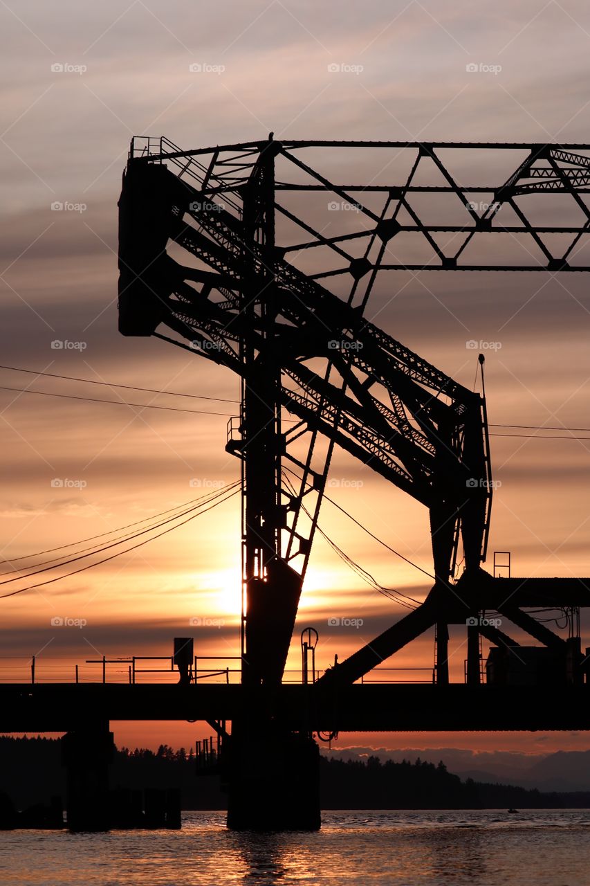 A deep orange sunset in the Pacific Northwest creates a dark silhouette on a bridge. The waters of the Puget Sound are seen in the backdrop.