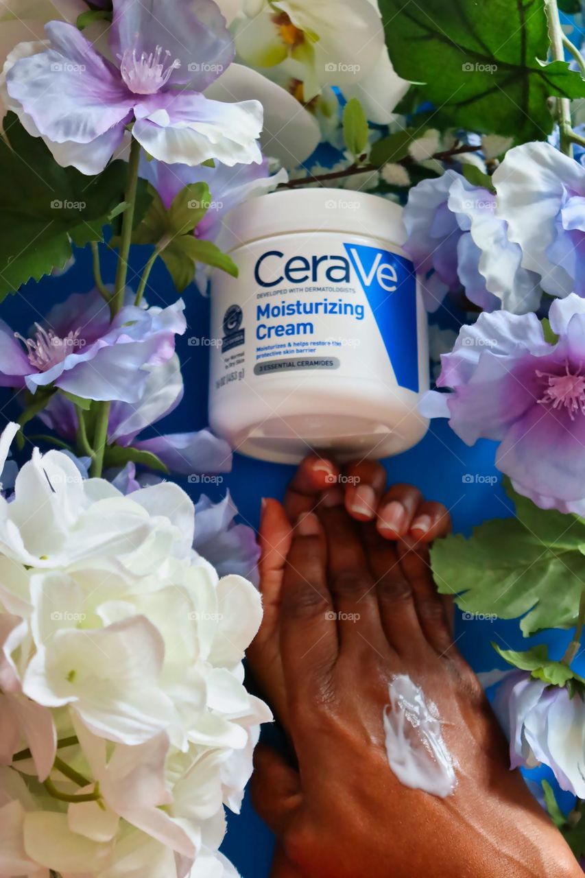 Woman applying Cerave Moisturizing Cream on blue background with flowers. 