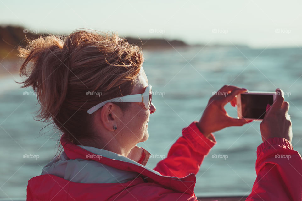 Young woman taking a photos using phone, looking at screen, standing outdoors, she is backlighted by sunlight with plain sky and sea in the background