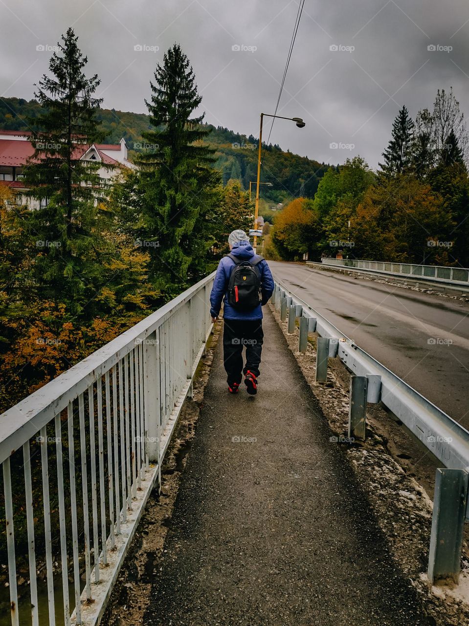 Man walking down the bridge road. Buildings rooftops among high green pines and colored leaves trees. Mountain covered with forest at the background. Walking man, tourist, travelling, exploring the world. Beautiful nature, pathway, the road far