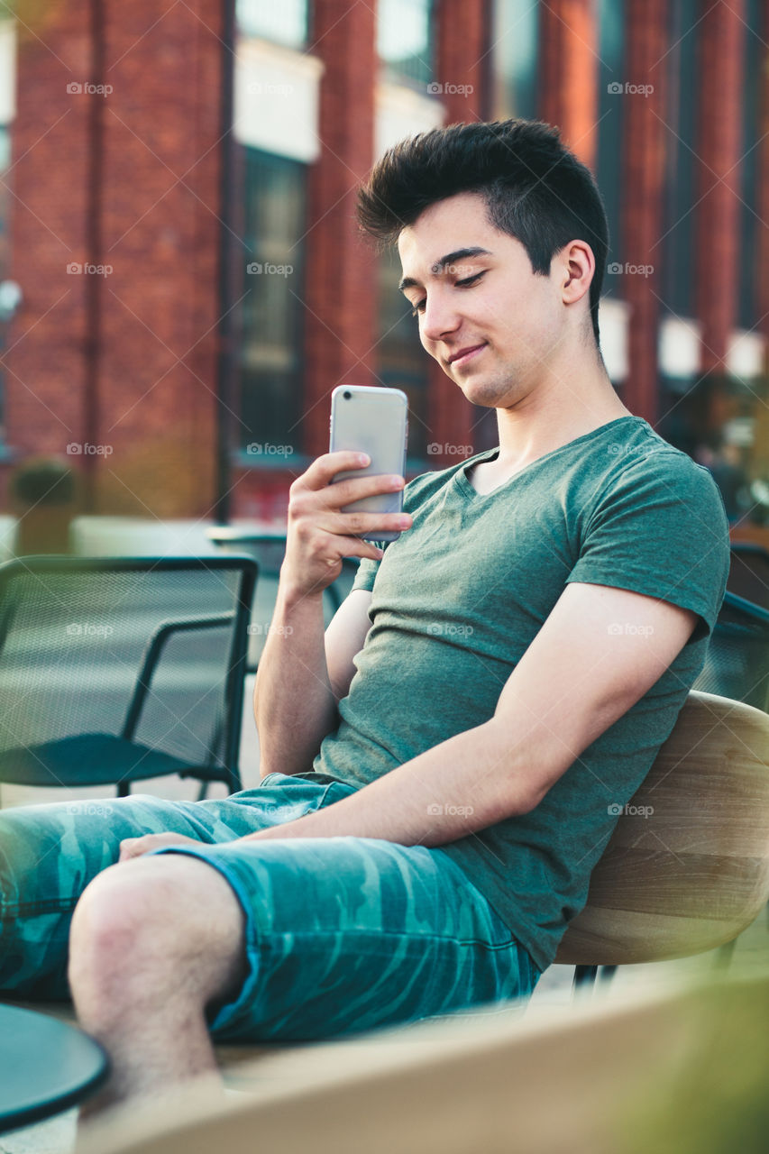 Young man having fun with smartphone, reading funny texts, sitting in center of town