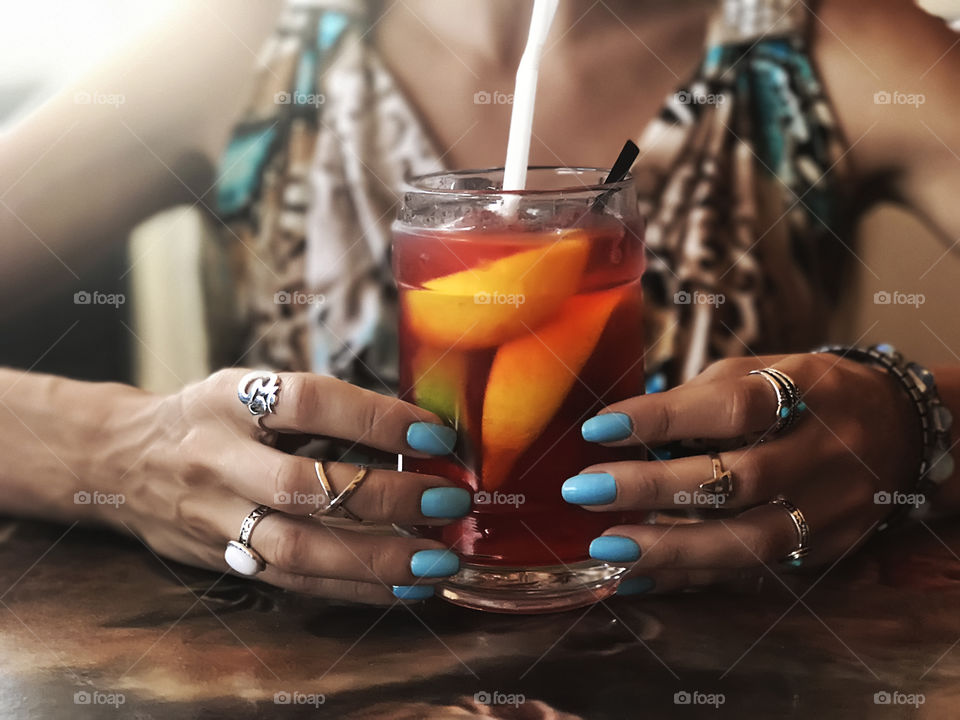 Young woman wearing fashionable rings with blue nail polish in colorful summer dress enjoying a tropical cocktail 
