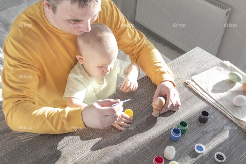 A little son and his father together in their free time paint Easter, chicken eggs with paints and brushes for the holiday.  A child and a man create a craft and natural product in their family business.