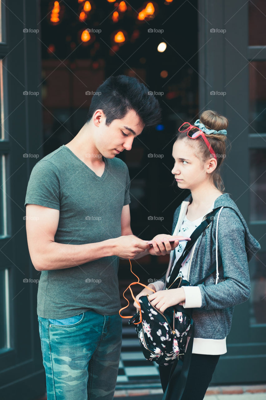 Couple of friends, teenage girl and boy,  using smartphones, talking together, standing on street in center of town, spending time together