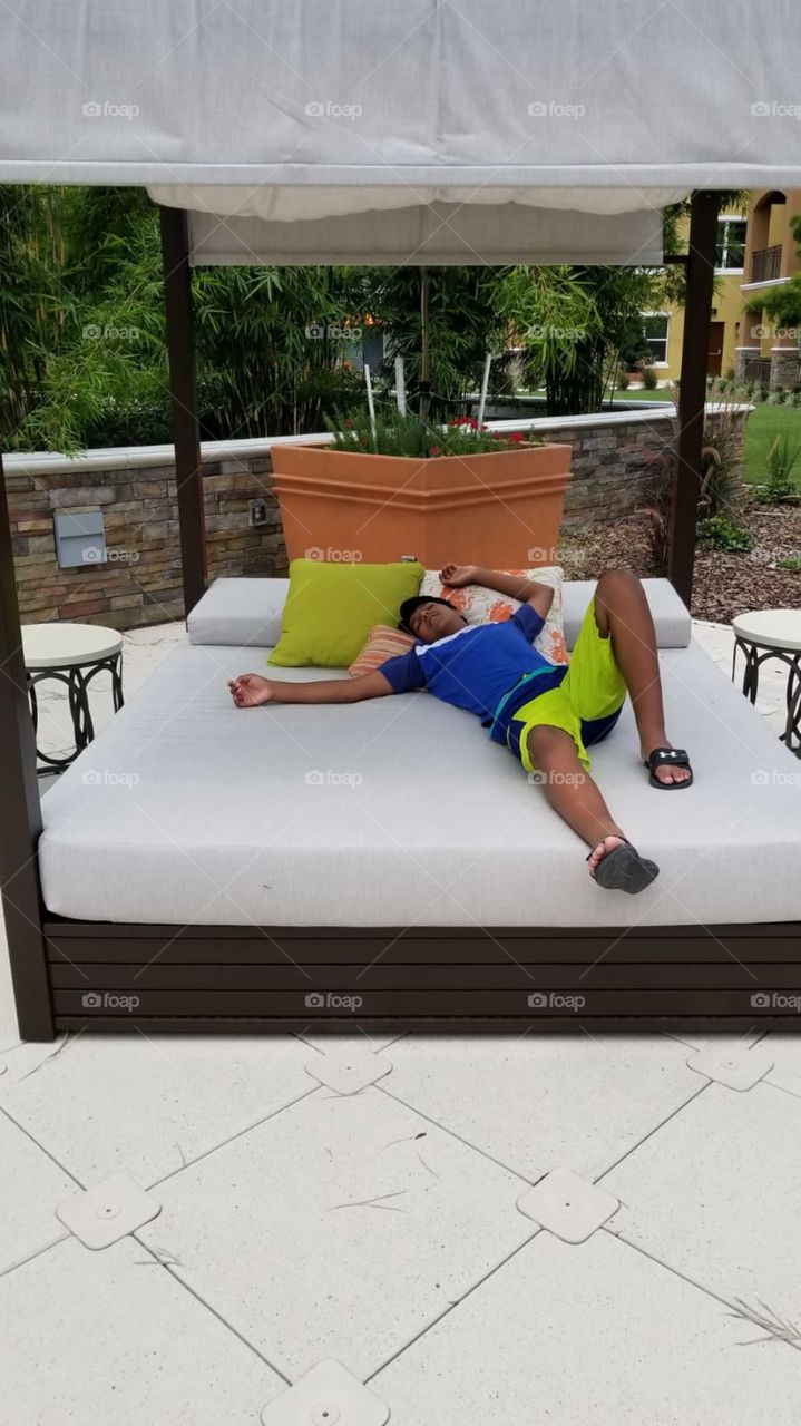 Rest and relaxation. Cabanaahhhh. Kid relaxing in pool cabana. Boy, bright colors, outdoors, swimming, calm, mattress, pillows, patio, furniture, evening, cabana