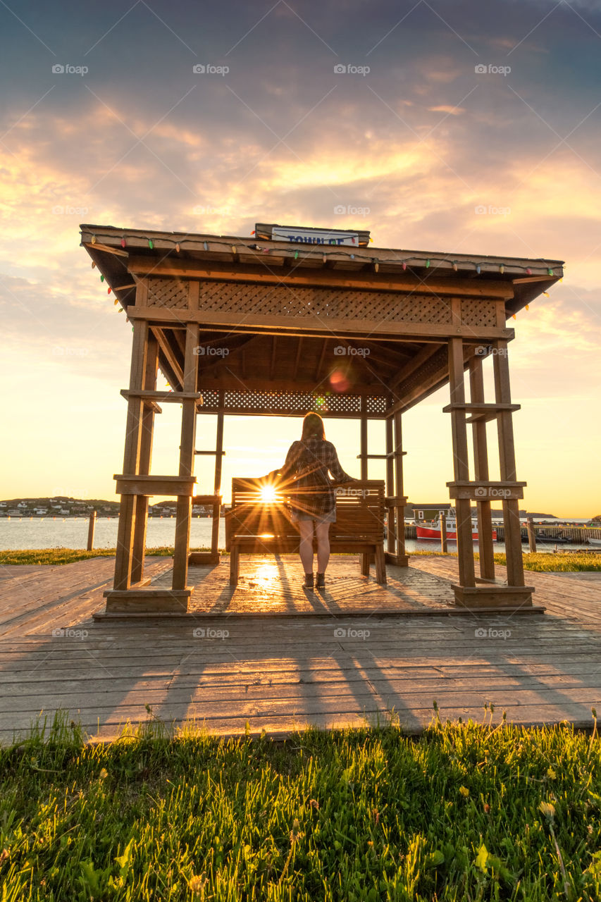 Young woman standing in a wooden gazebo watching the sun set. Sun star and flare through the bench