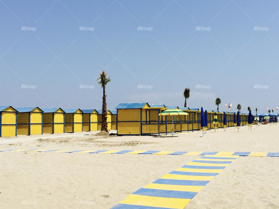 Italian sandy beach with yellow and blue beach cabins and palms with blue sky in the background 