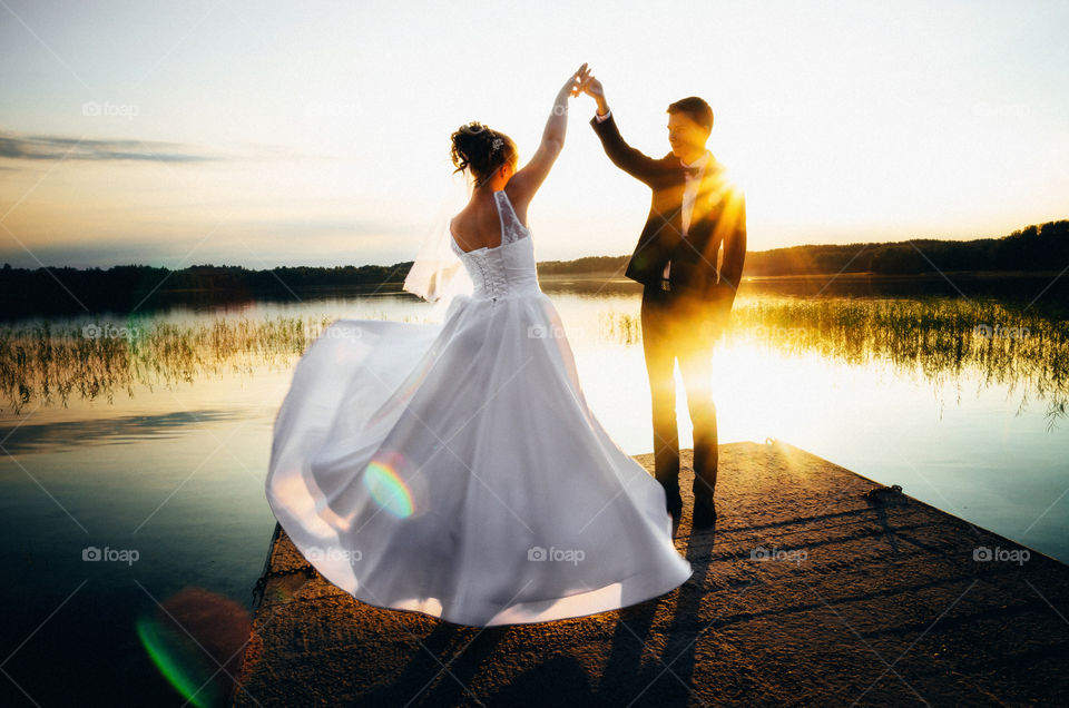 The groom holds bride's hand on the lake shore at sunset