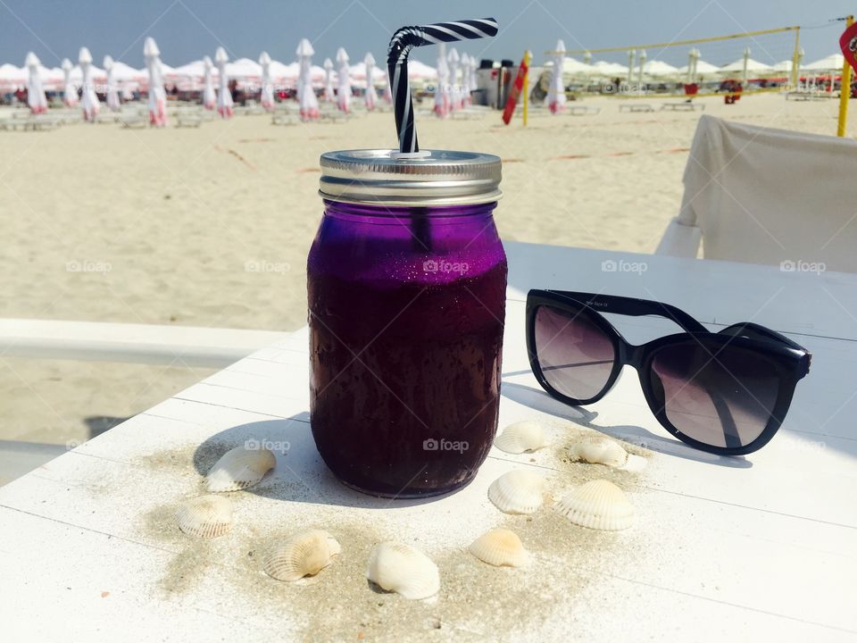 Jar of blueberries lemonade on white wooden table surrounded by seashells and pair of black sunglasses