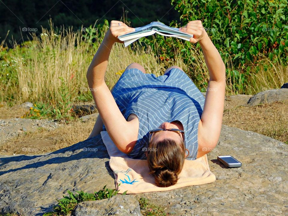 SUNNY DAYS - A young woman is lying on a rock, on her back, reading a book in the sun. Close up. Shadow. Concept of education