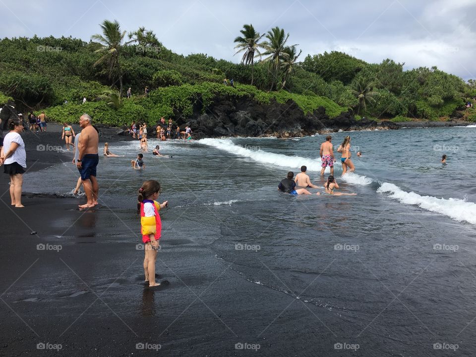 People frolicking on the black sand of a beautiful beach on the Road to Hana in Maui, Hawaii with the waves breaking and wind blowing the palm trees in the background.