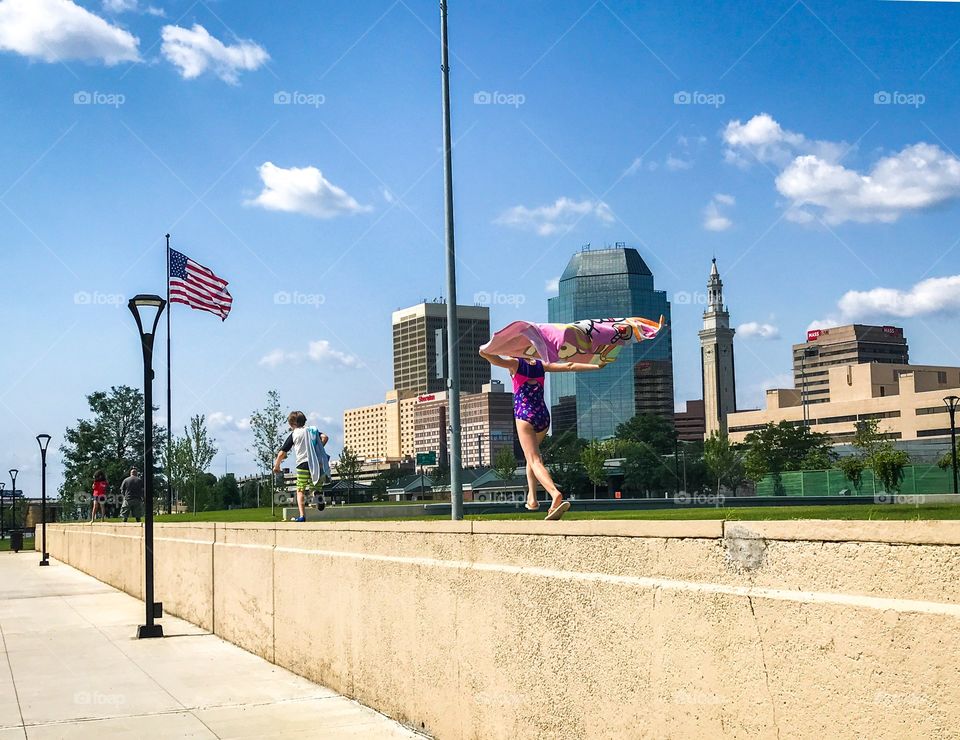 downtown care free children running with cape down cement wall in front of tall city buildings blue skies American flag 