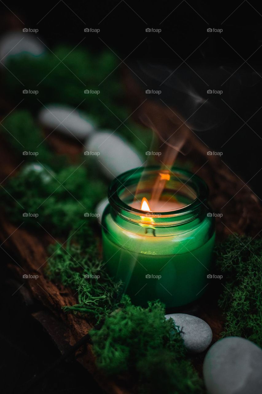 Beautiful view of a candle in a green glass jar standing in a natural tree bark with gray stones, moss and flowing smoke, close-up side view.