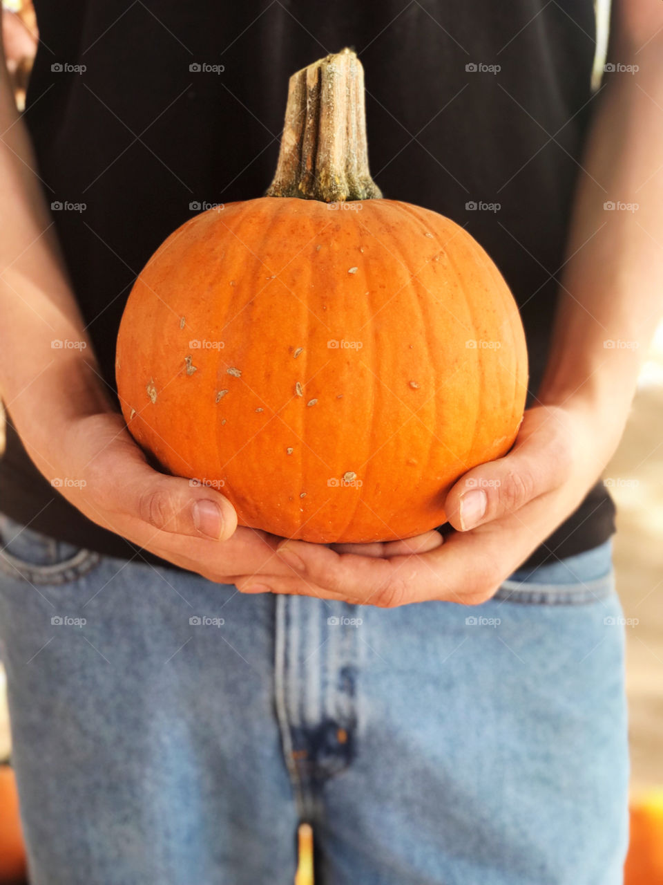 Male wearing black shirt and blue jeans holding a small pumpkin 