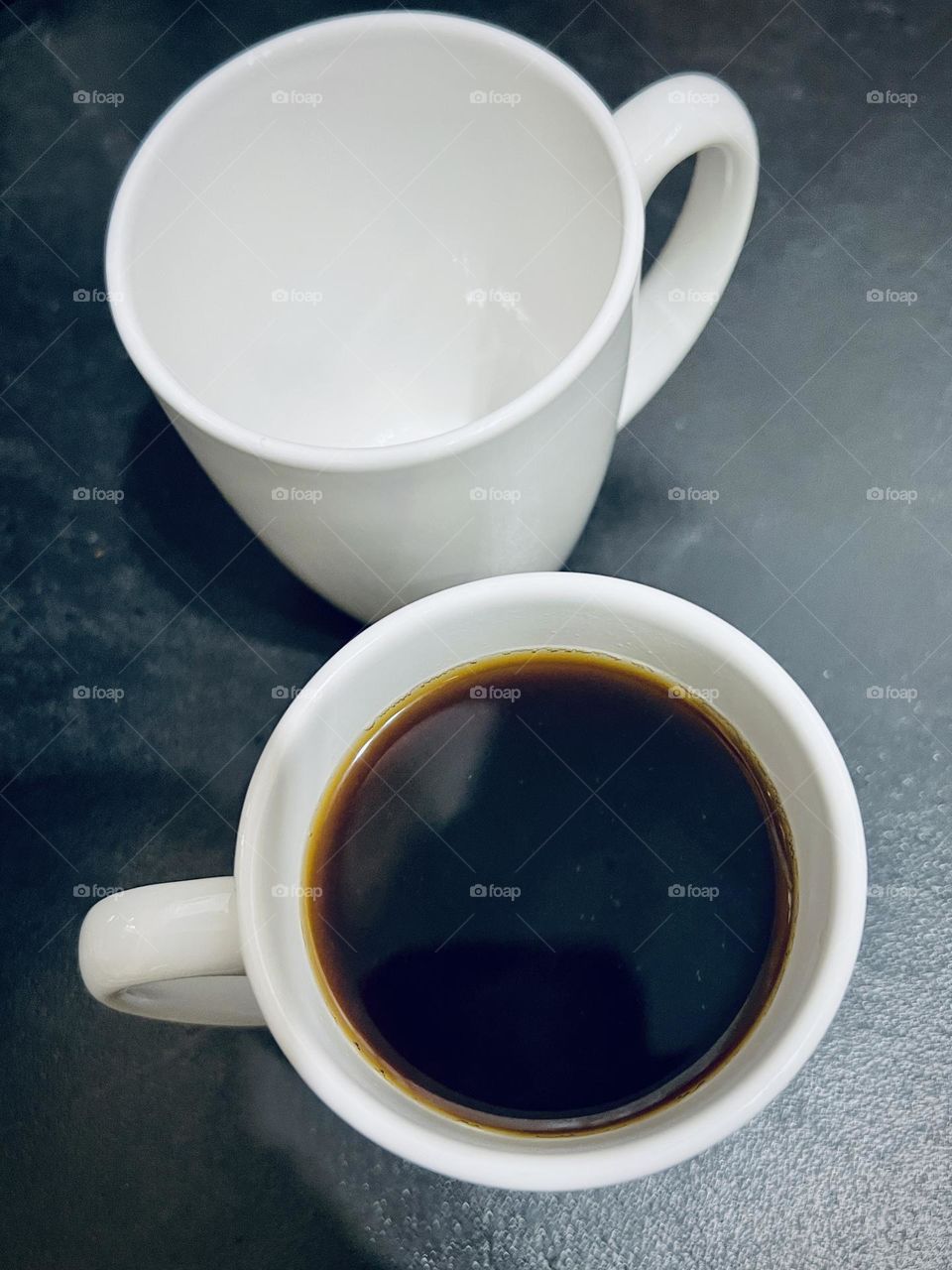 Overhead closeup of two white ceramic coffee cups. Cups are on a black laminate  countertop. One is full of black coffee while the other is empty.