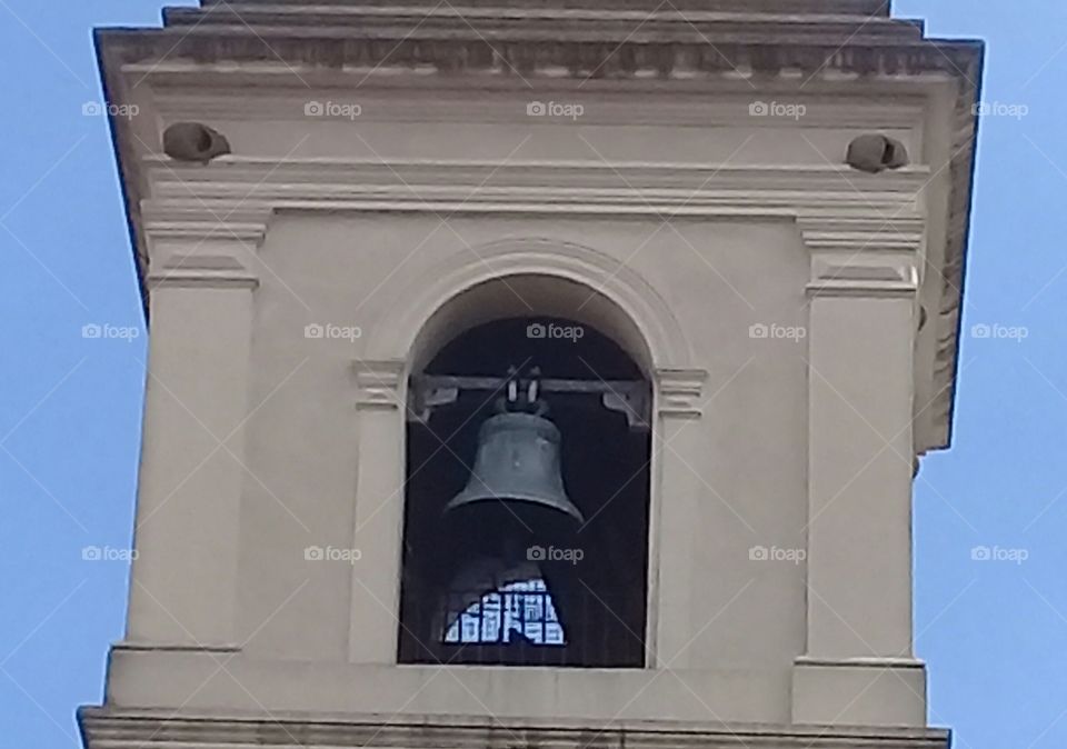 campana de torre de una antigua catedral, encerrada en una ventana con rejasy rodeada por una pared de color gris original.