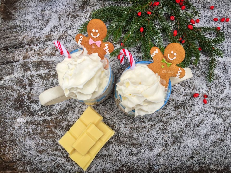 Two mugs of white chocolate with whipped cream, gingerbread man and candy canes on a snowy table with pieces of white chocolate and evergreen branches beside