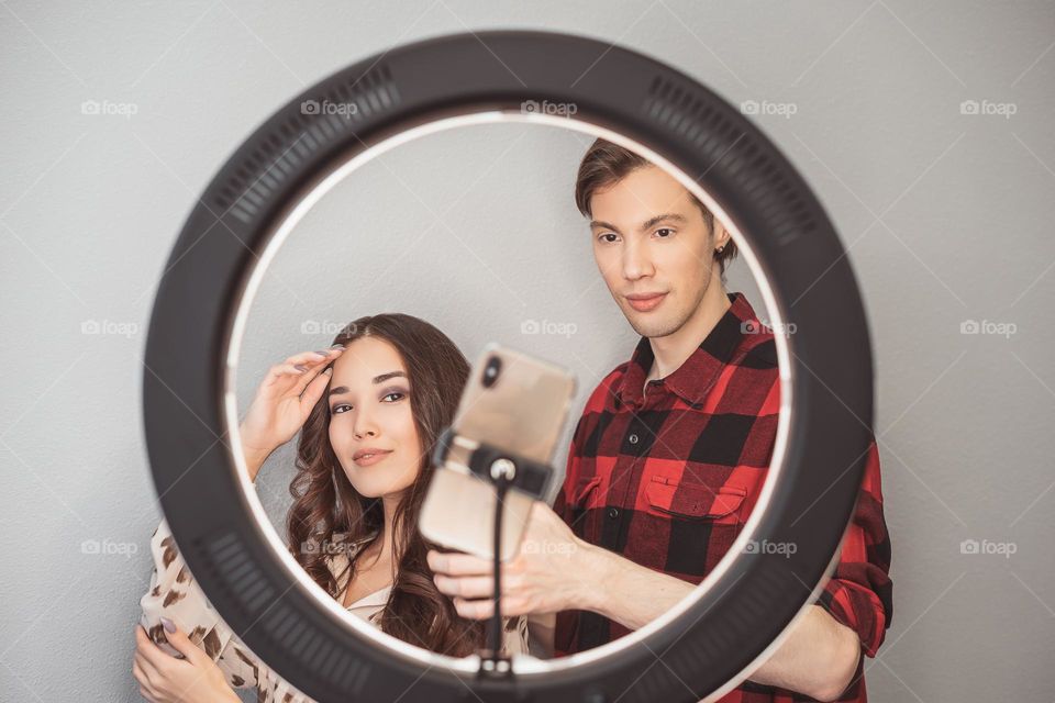 Young couple of beauty bloggers, hair stylist, hairdresser and model with hair style shoot on mobile phone with ring lamp on the grey wall background