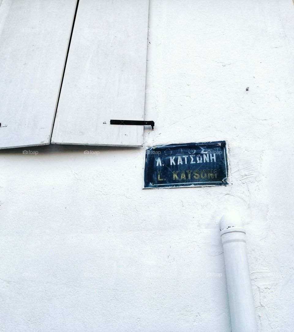 All white street wall in Greece, with a blue street sign on it and white window covers/ Simple white wall on the side of the street