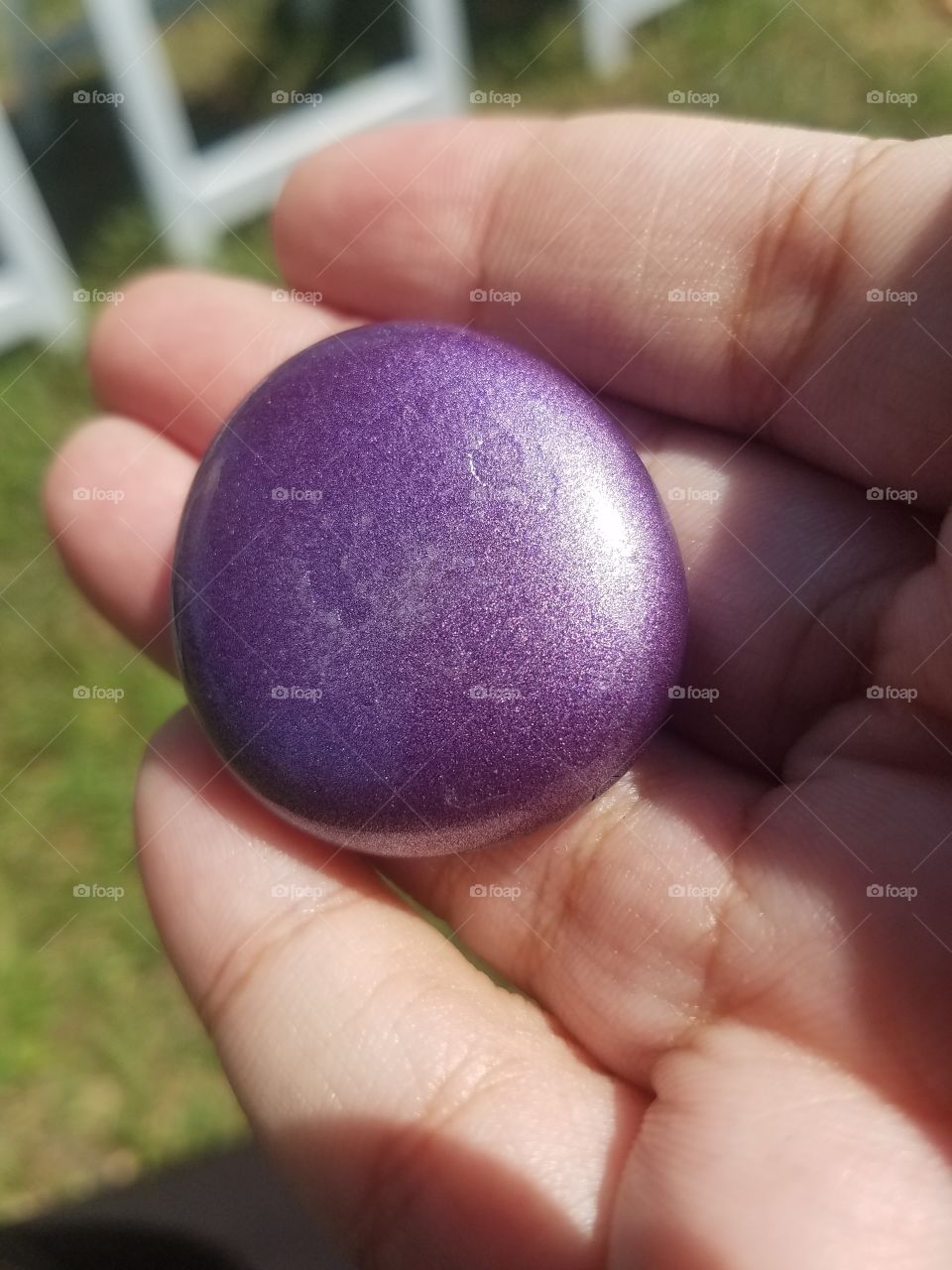 A hand holding a round, purple rock that is gleaming in the sun. The rock is fake.
