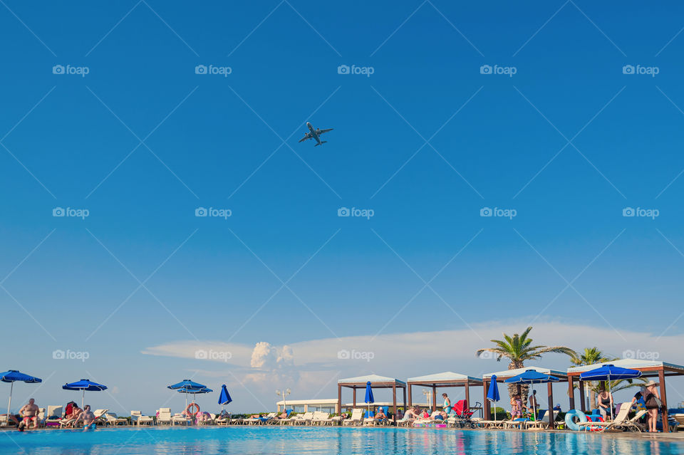 Airplane in the sky above tourists at the swimming pool in a holiday resort.