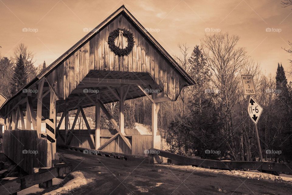A sepia photograph of a rustic covered bridge on a dirt road in the middle of winter in New England.