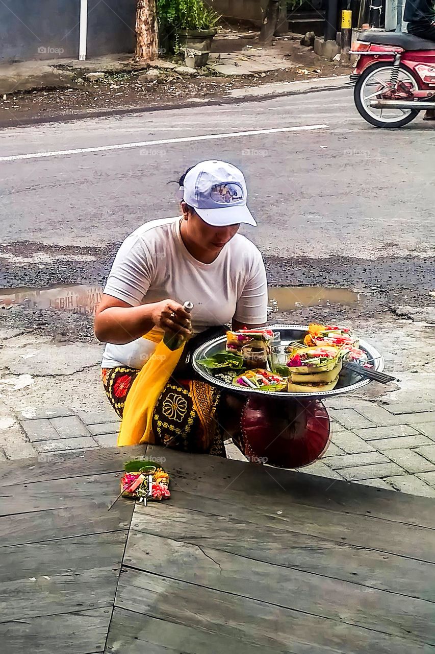 Denpasar, Bali, Indonesia - September 25, 2023: A woman wearing a hat and traditional clothing is placing offerings in front of a shop in Bali, Indonesia