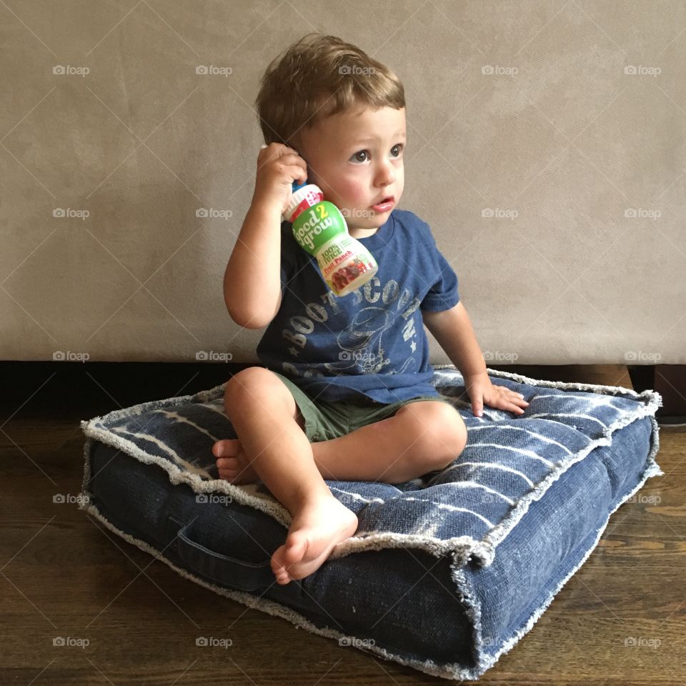 Toddler boy sitting on floor pillow talking on juice box phone. 