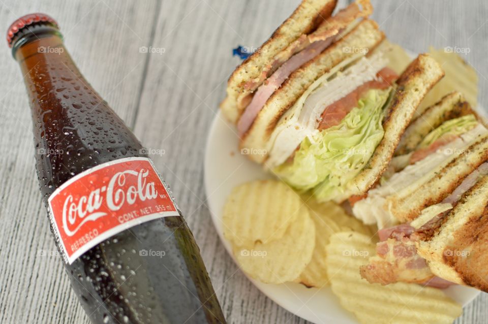 A can and a Bottle of Coca Cola with a club sandwich on a white rustic background 