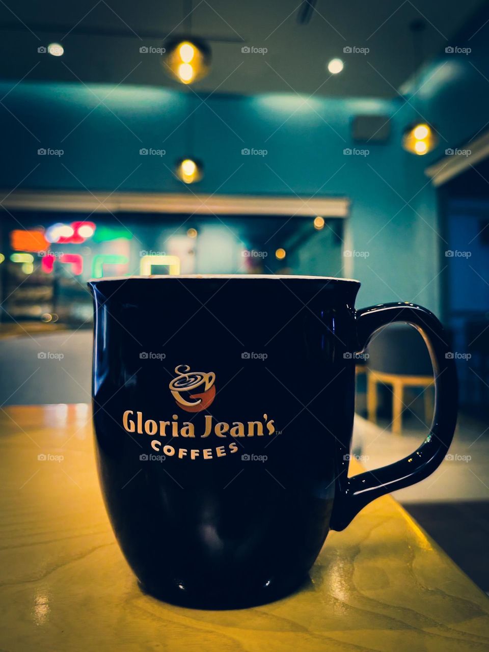 A black coffee mug, with Gloria Jean’s Coffees on it, at a Gloria Jean’s store. Soft ambience, predominantly green.