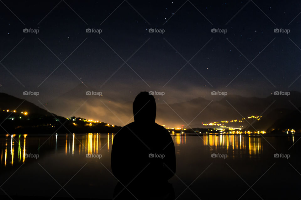 Peaceful picture of a guy looking at the calm scene in front of him. He looks at the reflection of the city lights on top of the lake.