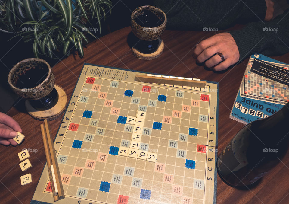 Wine & games! 2 people sit around an old and well used scrabble board, playing the game and drinking wine